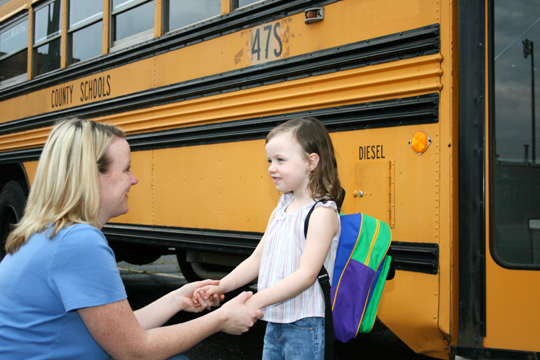 Image of a woman lovingly holding a small girl's hands in front of a school bus